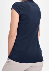 Person som bär en marinblå kortärmad skjorta och ljusbeige byxor, sedd bakifrån mot en enfärgad vit bakgrund.