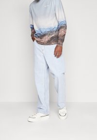 Helle blaue Denim-Hose mit lockerem Schnitt, kombiniert mit einem langärmeligen Shirt mit einem Farbverlauf eines Berglandschaftsdesigns und weißen Sneakers.