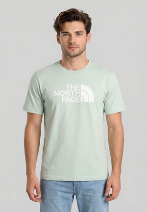 Joven con cabello castaño que lleva una camiseta de manga corta verde pálido con el logo blanco de "The North Face" y jeans azul claro.