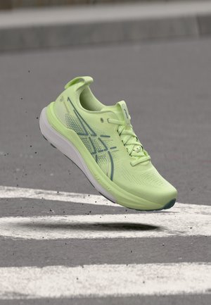 Chaussure de course vert clair avec semelle blanche, en l'air au-dessus d'une route goudronnée avec des marquages blancs, petites particules de saleté visibles autour de la chaussure.