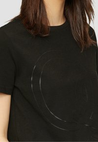 Svart bomullströja med en texturerad, glänsande logotypdesign framtill. Korta ärmar och rund halsringning. Mjuk tygkvalitet.