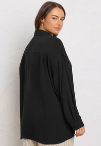 Camicia nera, oversize, con una leggera texture, maniche lunghe, polsini con bottoni e colletto classico, mostrata su un modello di spalle.