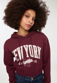 Bordowa, skrócona bluza z kapturem z białym napisem "NOWY JORK". Posiada sznurki i miękki materiał. Połączona z niebieskimi dżinsami.