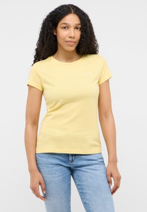 Frau mit langen lockigen Haaren, die ein hellgelbes kurzärmeliges T-Shirt und hellblaue Jeans trägt, steht vor einem einfarbigen Hintergrund.