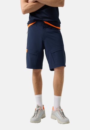 Mann trägt marineblaue Cargo-Shorts, weiße Socken und hellgraue Turnschuhe mit orangefarbenen Akzenten, steht mit verschränkten Armen.