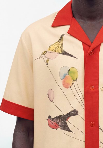 Chemise beige à manches courtes avec col et poignets rouges, ornée d'oiseaux imprimés et de ballons colorés suspendus à de fines ficelles.