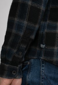 Zwart en blauw geruite langemouwen shirt met een gestructureerde stof en knoopmanchetten. Bevat een klein logo op de mouw. G gedragen met denim jeans.
