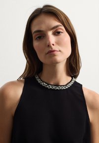 Top senza maniche di colore nero con collo alto, presenta un colletto decorato con perline in tonalità argentate. Tessuto dalla texture liscia, silhouette aderente.