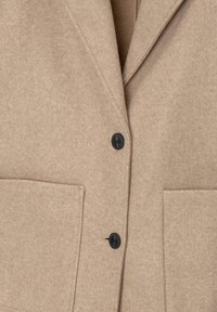 Manteau en laine beige avec deux boutons noirs, larges revers et grandes poches rectangulaires à l'avant.