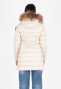 Crèmegekleurde pufferjack met een met bont afgewerkte capuchon, voorzien van gequiltte secties en een getailleerde pasvorm, gedragen over een blauwe jeans.