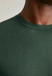 Olivegrünes T-Shirt mit Rundhalsausschnitt, aus strukturiertem Baumwollstoff gefertigt, hat ein tailliertes Design mit sichtbaren Nähten entlang des Ausschnitts.