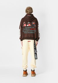 Joven de pie de espaldas, vestido con una sudadera marrón con texto motivacional y pantalones cargo crema con una correa gráfica negra adjunta.