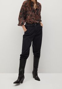 Blouse noire à motifs avec un col froncé, associée à un pantalon noir taille haute et des bottines en cuir à bout pointu avec une finition texturée.