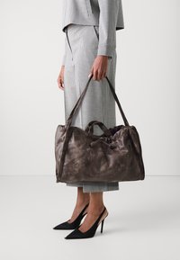 Sac de voyage en cuir marron avec une texture douce, fermeture à cordon et deux poignées robustes, présenté à côté d'un costume à carreaux et de talons noirs.