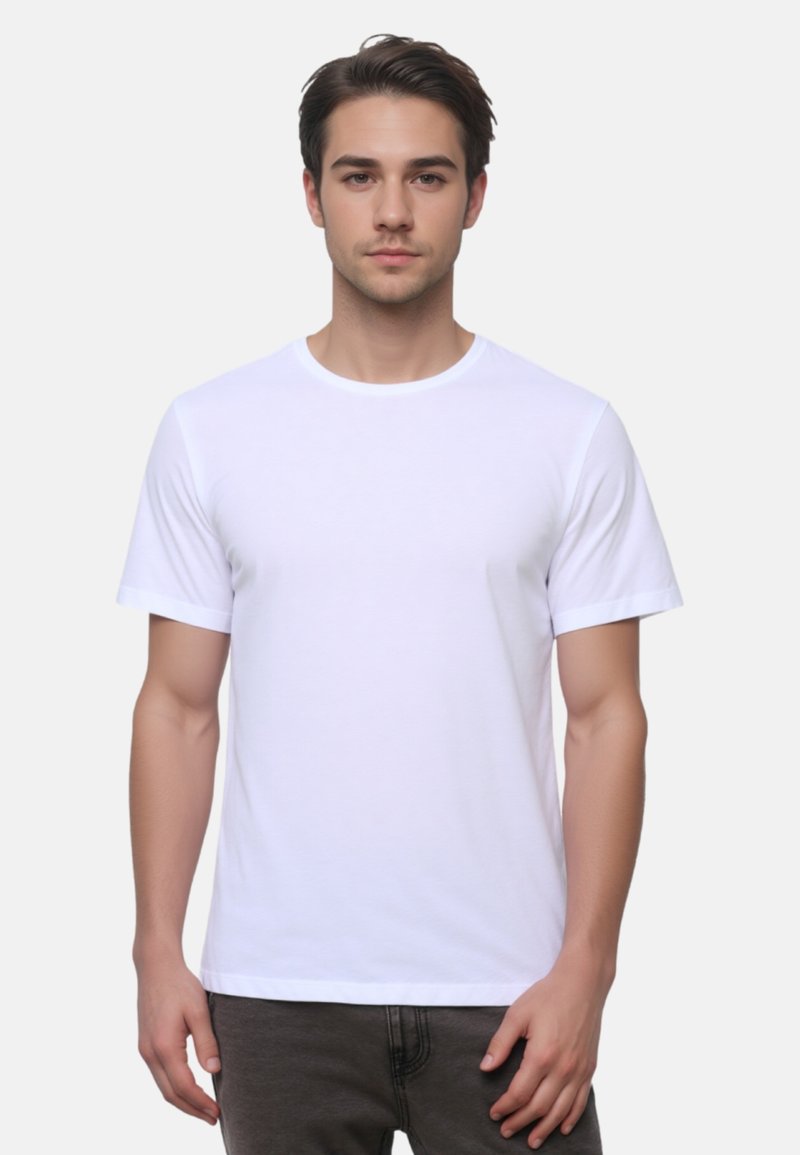 Junger Mann mit kurzen dunklen Haaren, der ein schlichtes weißes T-Shirt mit kurzen Ärmeln und dunkle Hosen trägt, vor einem hellgrauen Hintergrund steht.