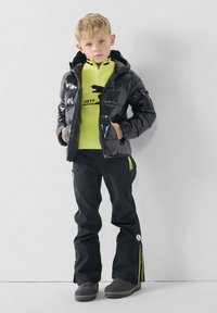 Chaqueta acolchada negra con textura brillante, camiseta intermedia amarilla con un logo, pantalones negros con detalles amarillos y botas grises.