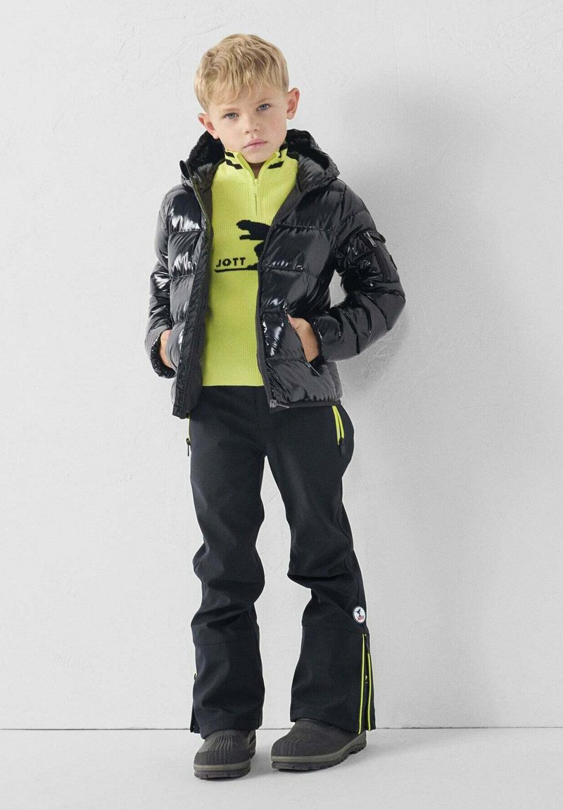 Chaqueta acolchada negra con textura brillante, camiseta intermedia amarilla con un logo, pantalones negros con detalles amarillos y botas grises.