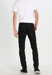 Černé slim-fit džíny s bezšvým designem a čtyřmi kapsami, kombinované s jednoduchým bílým tričkem a černými teniskami, stojící na světlém pozadí.
