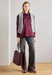 Chemise boutonnée bordeau, pull gris sur les épaules, jeans évasés gris, sac fourre-tout bourgogne et baskets marron. Fond en parquet.