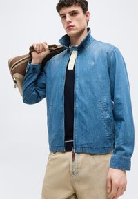 Νεαρός άνδρας φοράει μπλε τζιν μπουφάν πάνω από μπλε-ναυτικό και κρεμ πόλο μπλουζάκι, μπεζ παντελόνι, κρατώντας μια καφέ και μπεζ τσάντα ταξιδιού στον ώμο του.