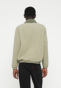 Άνδρας που κοιτάζει προς τα πίσω φορώντας ανοιχτό μπεζ μπουφάν με ελαστική κάτω ραφή και μανσέτες, συνδυασμένο με μαύρο τζιν, σε απλό λευκό φόντο.