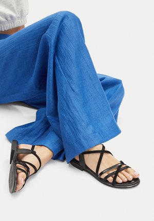 Schwarze Riemchensandalen mit flacher Sohle, ausgestattet mit überkreuzten Riemen und einer strukturierten Oberfläche, kombiniert mit hellblauen weiten Hosen.