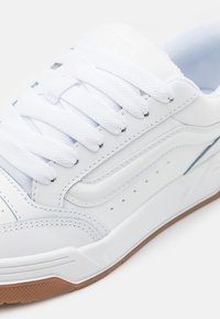 Weißer Sneaker mit glattem Lederobermaterial, gepolsterter Zunge, flachen weißen Schnürsenkeln, perforierten seitlichen Details und brauner Gummisohle.