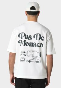 Camiseta blanca de gran tamaño con un gráfico negro de coches y el texto "Pas De Monaco" en la parte posterior; mangas cortas y escote redondeado.