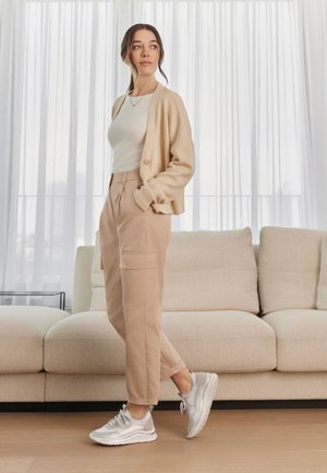Beige Strickjacke über einem weißen Oberteil, kombiniert mit hellbeigen Cargo-Hosen mit Seitentaschen und grauen Turnschuhen. Glatte Materialien, lässiges Design.