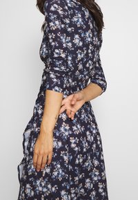 Robe florale navy avec un corsage ajusté et des manches longues. Présente une jupe évasée et des motifs floraux bleus et crème.