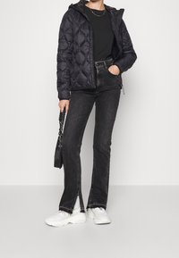 Chaqueta acolchada negra tipo puffer con capucha, combinada con jeans negros de campana con aberturas laterales, zapatillas blancas y un pequeño bolso negro.