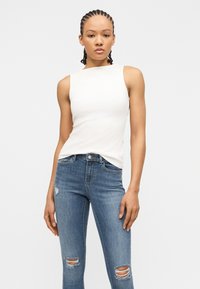 Νεαρή γυναίκα με πλεξούδες μαλλιά, φορώντας ένα λευκό αμάνικο τοπ και σκισμένο μπλε τζιν, στέκεται μπροστά σε ένα απλό ανοιχτόχρωμο φόντο.