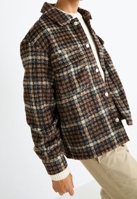 Osoba w brązowo-beżowej wełnianej kurtce w kratę z napami, kremowym swetrze i beżowych spodniach na tle białego tła.