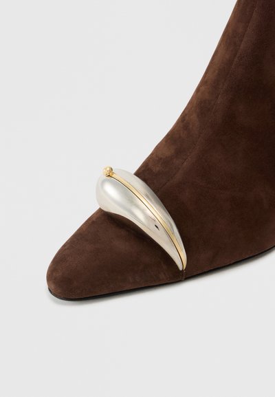 Brown suede heeled shoe with a pointed toe, featuring a silver-accented sculptural detail on the vamp and a gold trim.