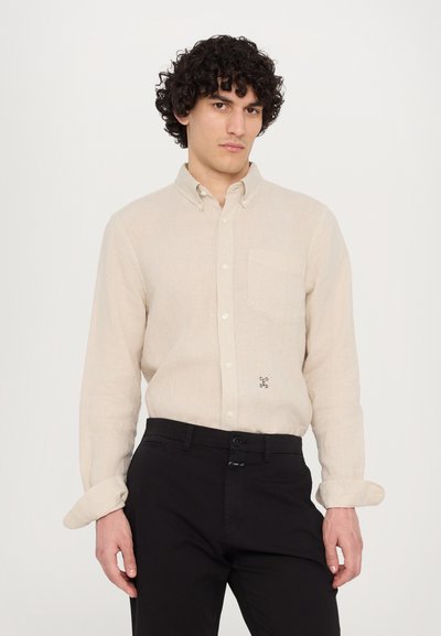 Chemise boutonnée en lin beige clair avec une poche poitrine et un détail de logo subtil. Associée à un pantalon foncé ajusté, mettant en valeur les manches retroussées.