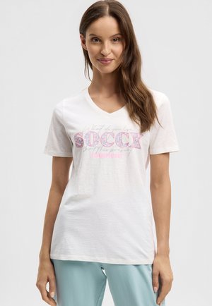 Junge Frau mit langen braunen Haaren, die ein weißes V-Ausschnitt-T-Shirt mit pinkem und blauem "SOCCX"-Schriftzug sowie hellblaue Hose trägt, vor einem schlichten Hintergrund.