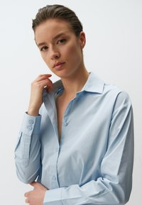 Camicia azzurra chiara con colletto classico. Presenta maniche lunghe, dettagli sui polsi e una texture liscia. Nessun motivo o decorazione.