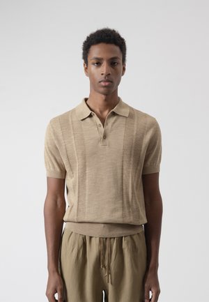 Joven con una camisa polo de punto beige de manga corta y pantalones caqui holgados, de pie frente a un fondo liso de color gris claro.