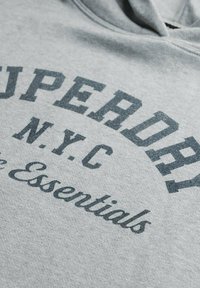Sudadera gris confeccionada en tela suave que presenta un atrevido texto negro que dice "SUPERDRY N.Y.C. Essentials". Cuello redondeado y diseño frontal.