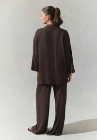 Braunes, strukturiertes, locker sitzendes Outfit, bestehend aus einem Langarmoberteil und weit geschnittenen Hosen, kombiniert mit schwarzen Sandalen mit offener Zehe und goldenen Accessoires.