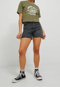 Camiseta corta de color verde oliva con estampado gráfico blanco, combinada con pantalones cortos de mezclilla negra con dobladillo deshilachado. Zapatillas altas negras.