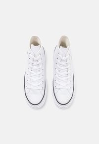 Baskets montantes blanches en toile, avec des lacets plats blancs, une toe ronde et une semelle en caoutchouc noire avec une bande blanche contrastante.