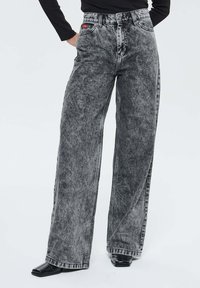 Femme portant un jean gris délavé à taille haute et jambes larges, avec des chaussures noires à bout carré et un haut noir à manches longues, se tenant avec une main dans sa poche.