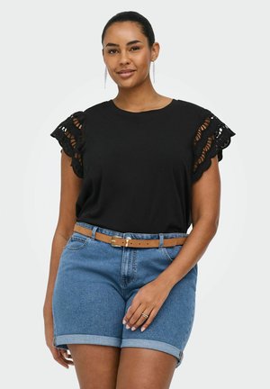 Woman wearing black short-sleeve top with lace ruffle sleeves, blue rolled denim shorts, tan belt, and dangling earrings, standing with relaxed posture.