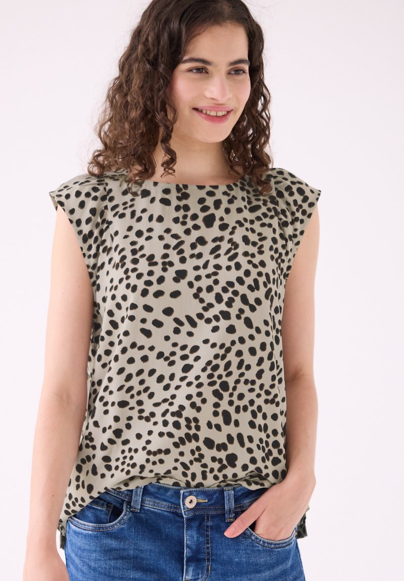 Femme aux cheveux bouclés souriante, portant un haut beige sans manches à pois noirs et un jean bleu, une main dans la poche.