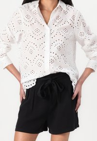 Blusa bianca in cotone con motivo a occhio ricamato, dotata di colletto e chiusura a bottoni. Abbinata a pantaloni corti neri con cordoncino.