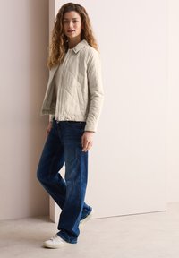Veste matelassée beige clair avec col, fermeture éclair, associée à un jean bleu foncé à jambes larges et des baskets blanches. Texture lisse, design décontracté.