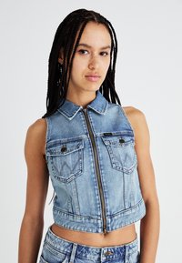 Giacca cropped senza maniche in denim azzurro chiaro con colletto, zip frontale e due tasche sul petto con cuciture decorative. Texture liscia.