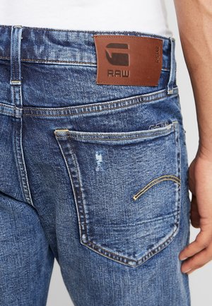 Vue arrière d'un jean en denim bleu avec des surpiqûres jaunes, un patch de marque en cuir et une main reposant près de la hanche droite.