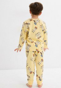 Bambino in piedi a piedi nudi, di spalle, che indossa un pigiama giallo con un motivo di suricati giocosi e un cappello blu e arancione.